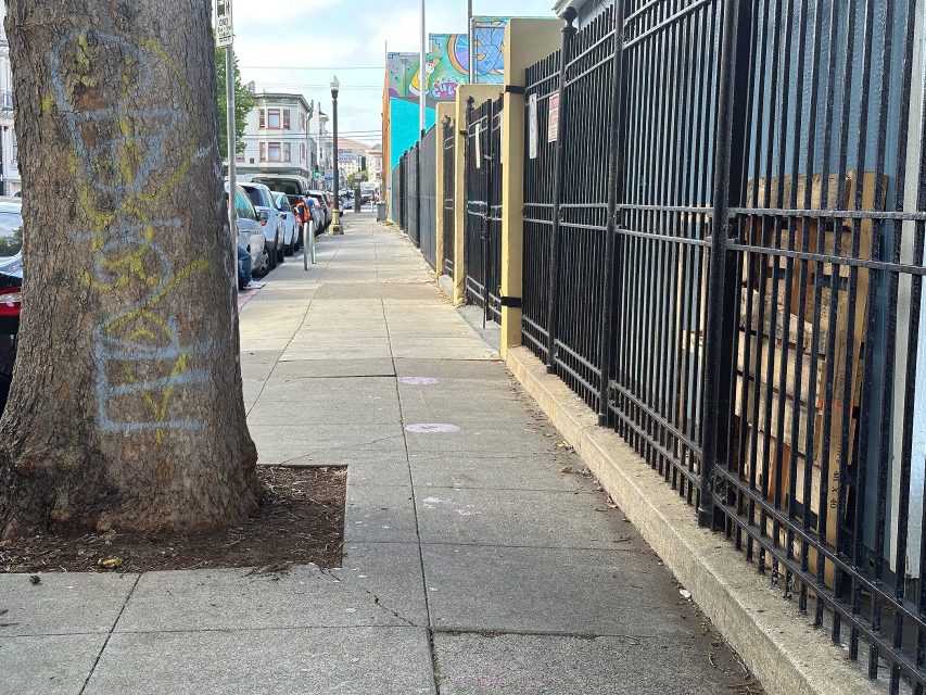 A sidewalk lined with parked cars on the left, a tree with chalk markings, and a black metal fence on the right; buildings and a colorful mural are visible in the background.