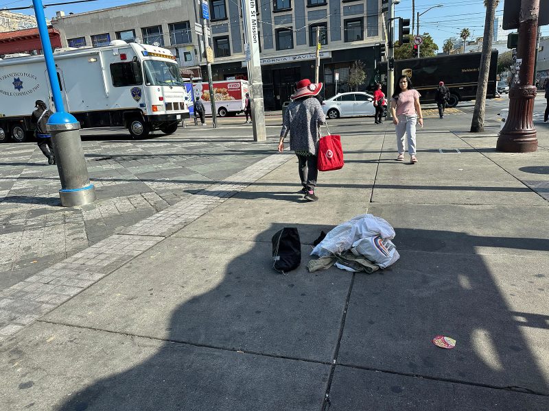 A woman in a red hat walks away from a pile of clothes on a city sidewalk; other pedestrians and a garbage truck are in the background.