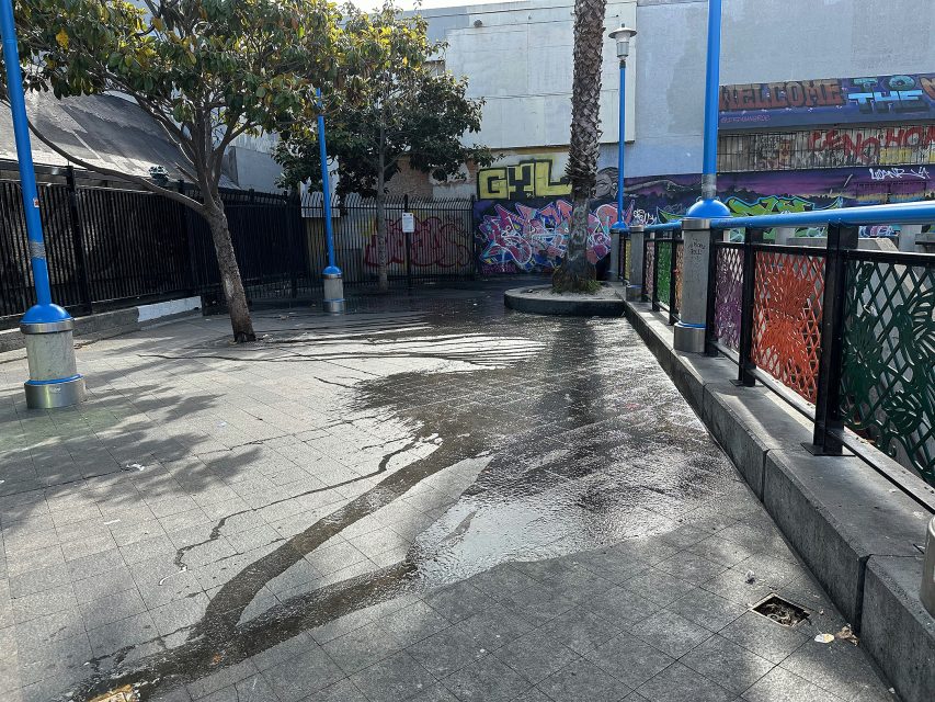 A wet urban plaza with puddles on the ground, surrounded by trees, blue lamp posts, a palm tree, and graffiti-covered walls.