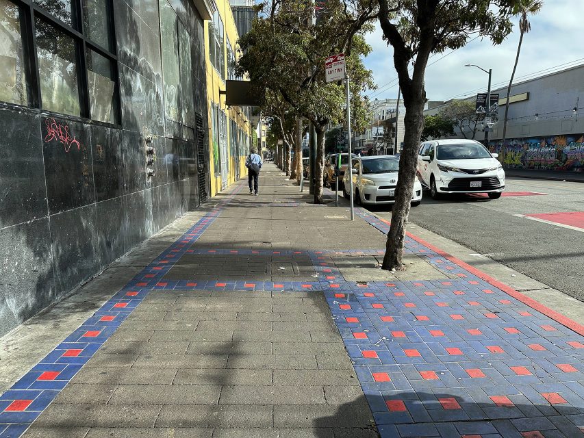 A city sidewalk with a blue and red tile border, trees, parked cars, and a person walking in the distance on a sunny day.