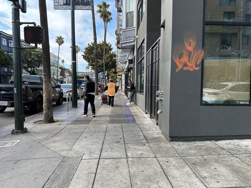 A city sidewalk with pedestrians, a worker in an orange vest pushing a cart, cars parked on the street, and orange graffiti on a building wall.