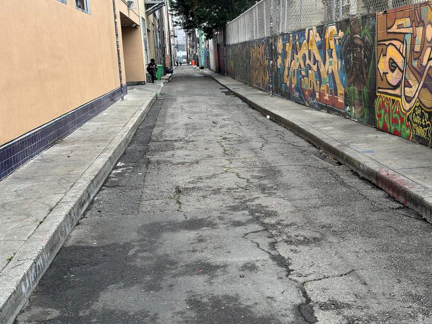A narrow urban alley with cracked pavement, graffiti-covered wall on the right, beige building on the left, and two people standing in the distance.
