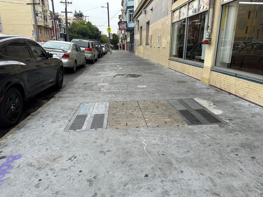 A city sidewalk with metal grates and utility covers, bordered by parked cars on the left and buildings with storefront windows on the right.