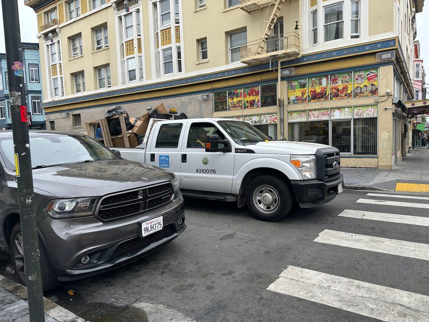 A gray SUV and a white city utility truck are parked at a street corner in front of a beige building with colorful window displays.