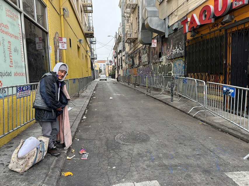 A person stands on an urban alleyway with graffiti and barricades, holding a blanket beside a bag with belongings near a closed shop.