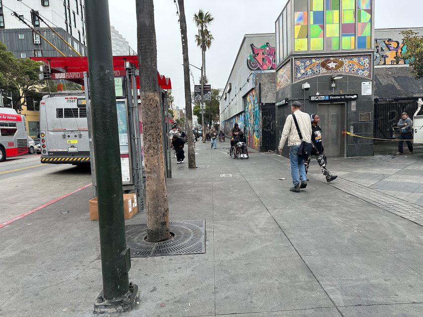 City street scene with people walking, a bus at a stop, colorful murals on building walls, and a palm tree lining the sidewalk.