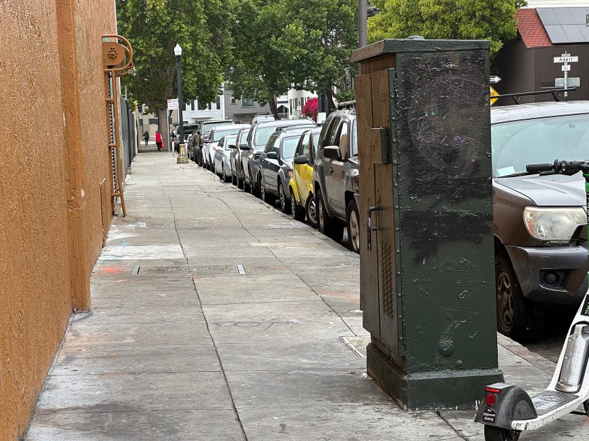 A sidewalk scene with parked cars, a green utility box, a scooter, and trees in the background on an overcast day.