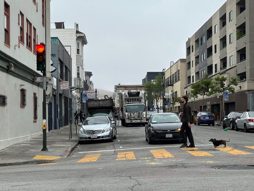 A man walks a small dog across a crosswalk at an intersection with a red traffic light; parked cars and a garbage truck are visible on the street.