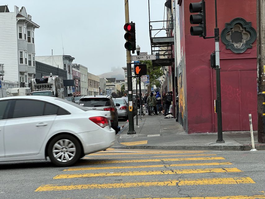 A white car drives through a crosswalk at a city intersection with a red light overhead and pedestrians waiting to cross near 16th Street.