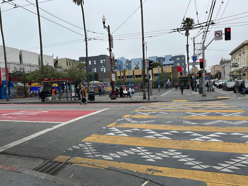 Urban street intersection with patterned crosswalks, people waiting at a bus stop, traffic lights, and buildings in the background on a cloudy day.