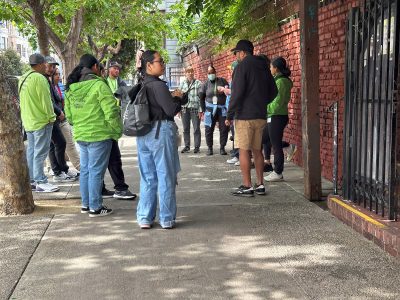 A group of people stand and talk on a city sidewalk next to a brick wall and trees, some wearing green jackets and casual clothing.