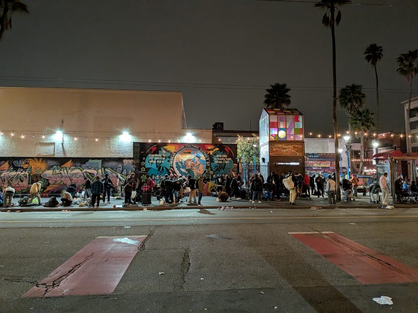 A large group of people gathers at night on a city sidewalk in front of a mural-covered building, under streetlights and string lights.