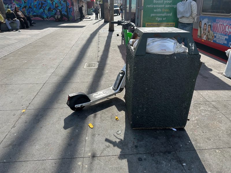 An electric scooter is tipped over, leaning against a trash bin on a city sidewalk in daylight; litter and people are visible in the background.