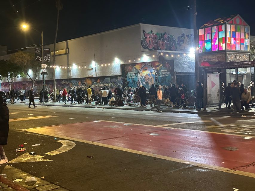 A large group of people gathers on a city sidewalk at night near a bus stop and a building with graffiti and murals, under streetlights and string lights.