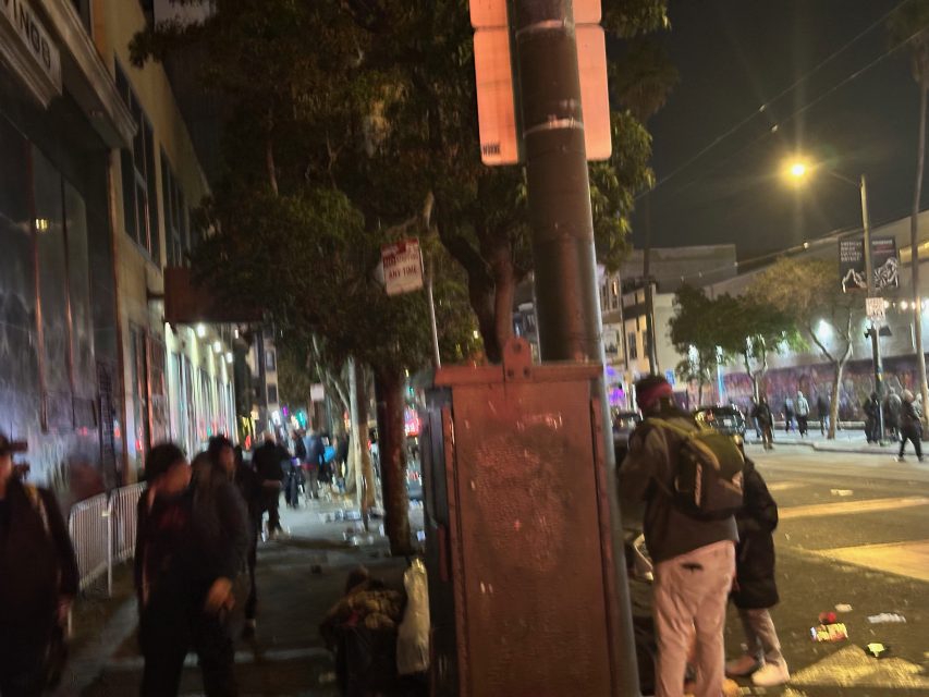 A city street at night with several people walking and standing near a sidewalk lined with litter, illuminated by streetlights and passing cars.