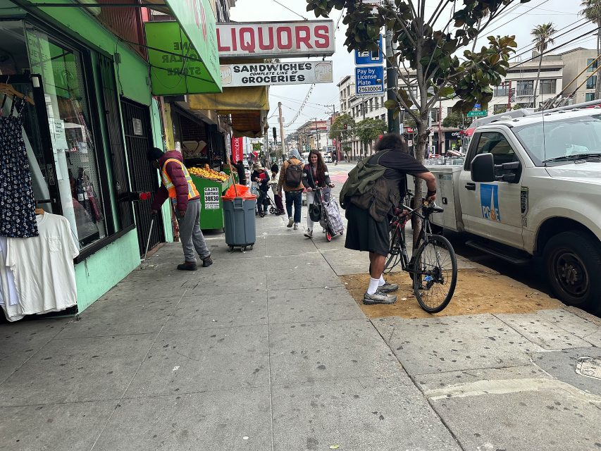 A man with a loaded bicycle stands on the sidewalk near a liquor store; pedestrians walk past shops and street signs in an urban setting.