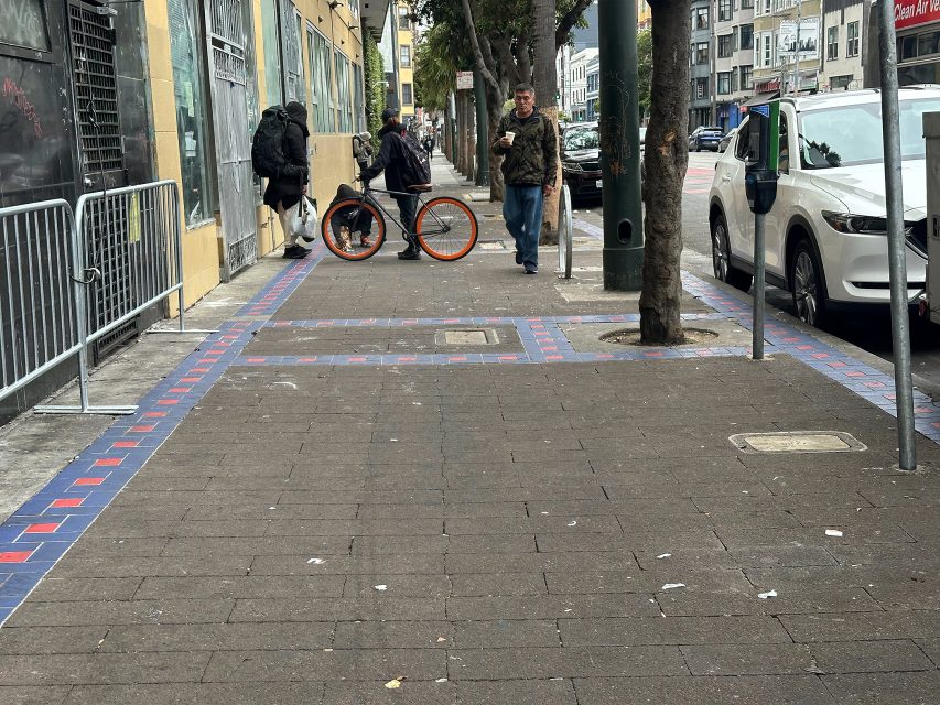 A city sidewalk with people walking, a person standing near an orange bike, parked cars, and buildings along the street.