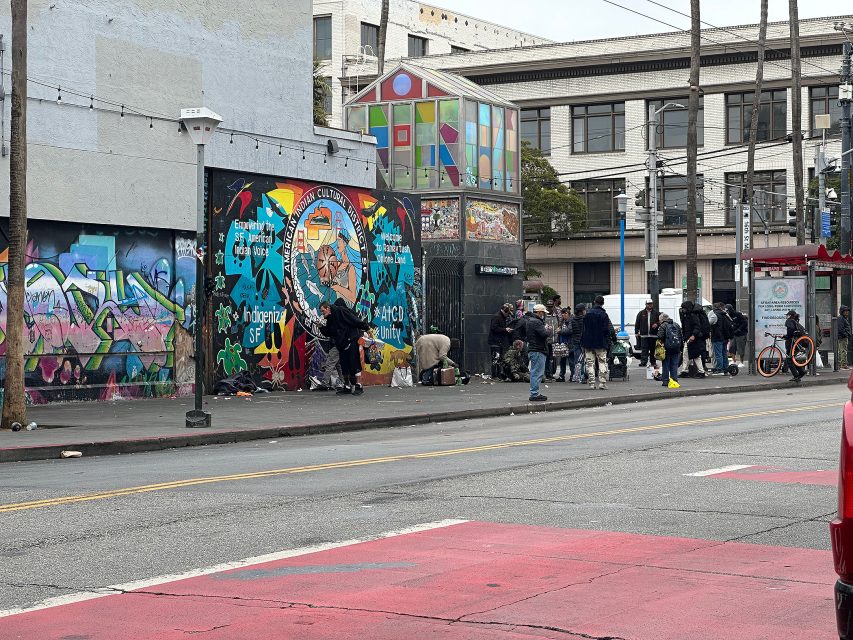 A group of people gather on a city sidewalk near colorful murals and graffiti, with scattered belongings and litter visible on the ground.