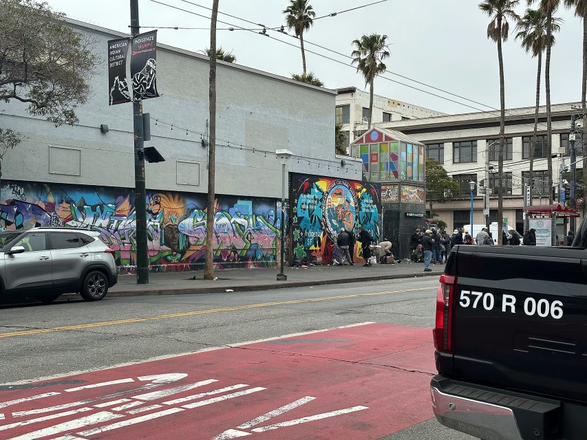 Street scene with a group of people gathered near a colorful mural, parked cars, and palm trees lining the sidewalk on an overcast day.