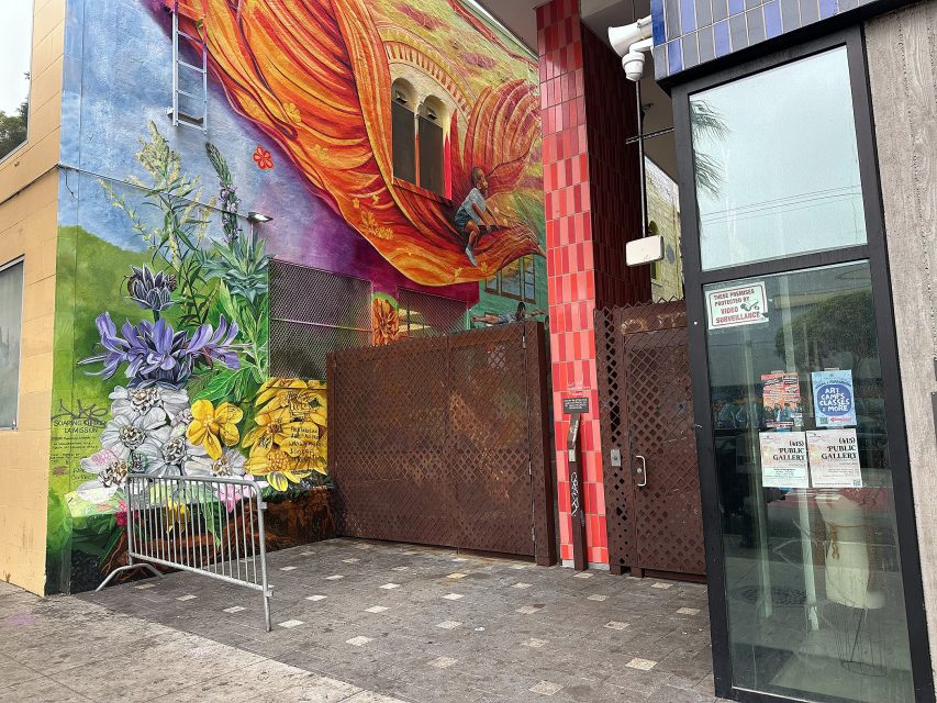 A colorful mural featuring flowers and a person climbing draped fabric covers a building wall next to a metal gate and a glass door with posted signs.
