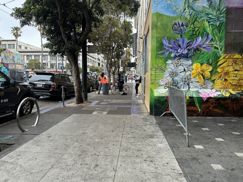 A city sidewalk with parked cars, people walking, a colorful floral mural on a building, and trees lining the street.