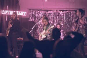 A band performs on stage with a saxophonist, guitarist, and drummer; a neon sign reads "OFTEN EASY" in the background, audience members are visible in the foreground.