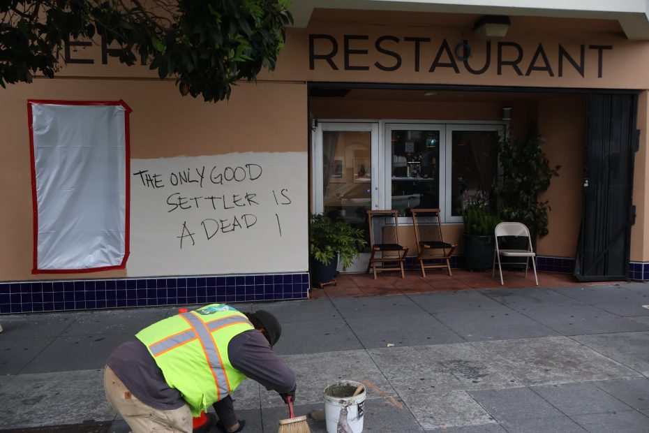 A person in a safety vest works outside a restaurant with graffiti that reads "THE ONLY GOOD SETTLER IS A DEAD 1" on the wall and a window covered in red-taped plastic.