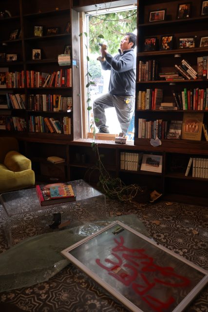 A person repairs a broken window in a library or bookstore, with shattered glass and a graffiti-covered window frame on the floor.