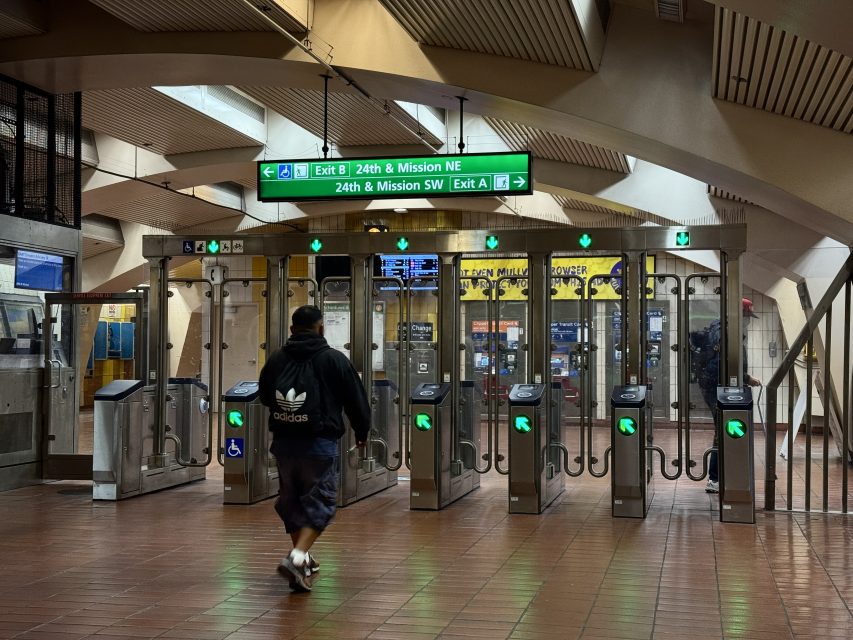 A person walks toward turnstiles at a subway station, with exit signs overhead showing directions for 24th & Mission NE and SW.