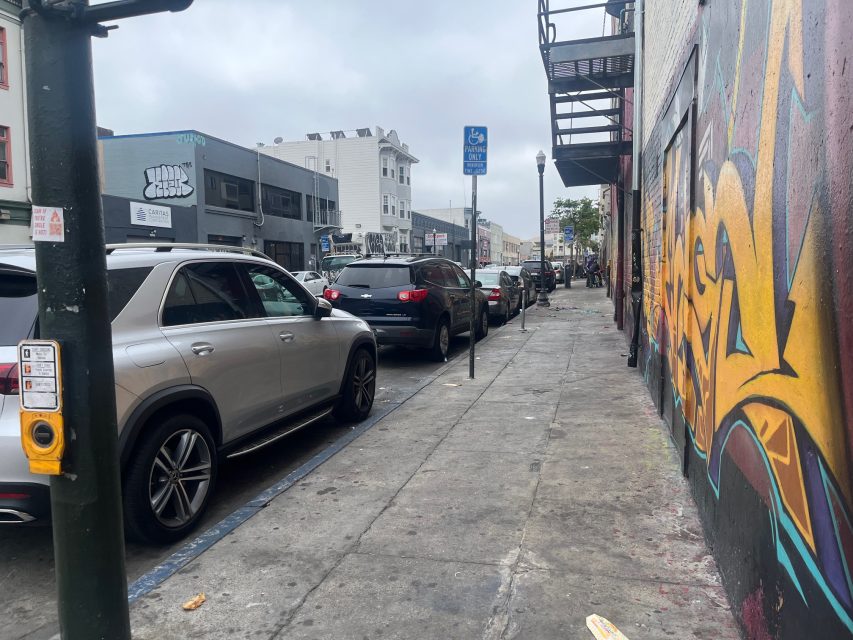 Street lined with parked cars, a graffiti-covered wall on the right, and a pedestrian crossing button on a pole in the foreground under a cloudy sky.