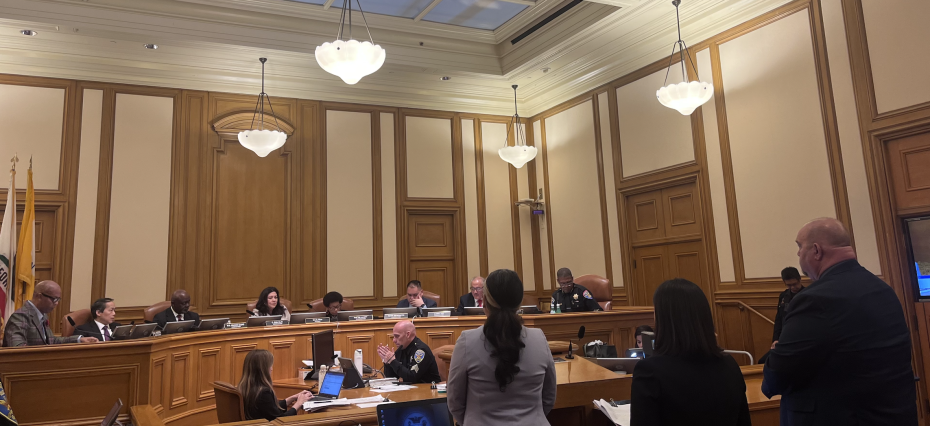 A city council meeting in session with officials seated at a long, elevated desk and attendees standing and sitting in front of them in a wood-paneled chamber.