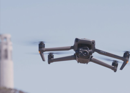 A quadcopter drone flies outdoors with blurred buildings and trees visible in the background.