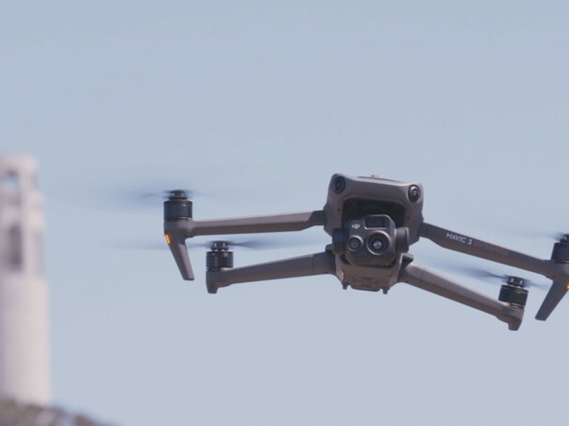 A quadcopter drone flies outdoors with blurred buildings and trees visible in the background.