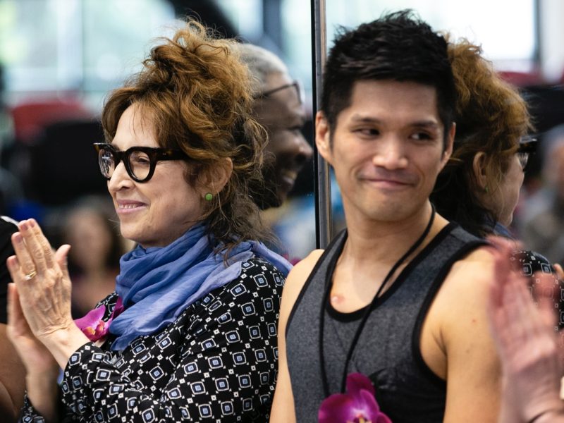 A woman with glasses and a blue scarf claps while standing next to a man in a sleeveless shirt; both wear flower leis, with a mirrored surface behind them.