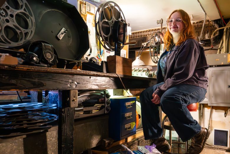 A person with long red hair sits on a stool in a workshop surrounded by film reels, equipment, and tools on a cluttered workbench.