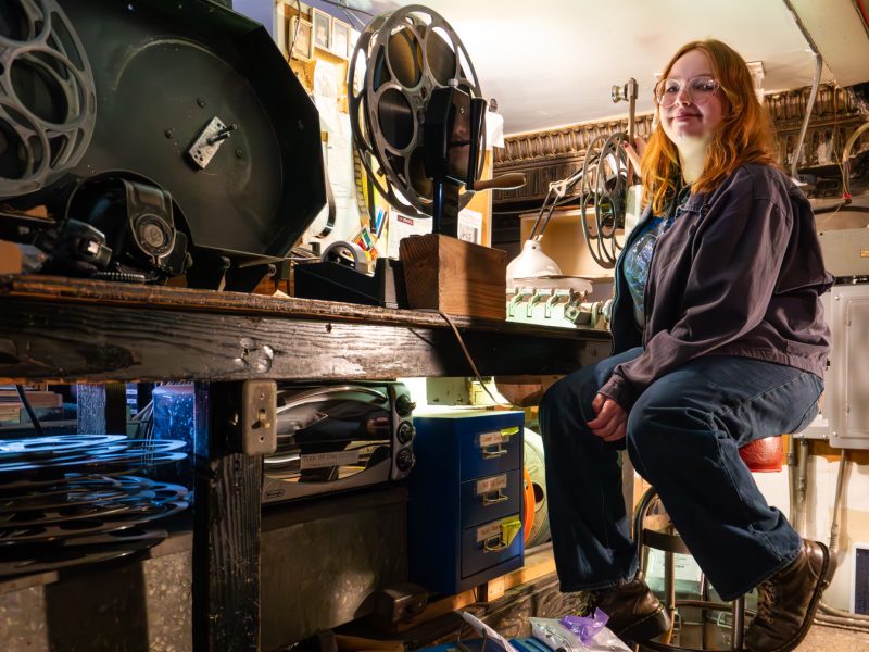 A person with long red hair sits on a stool in a workshop surrounded by film reels, equipment, and tools on a cluttered workbench.