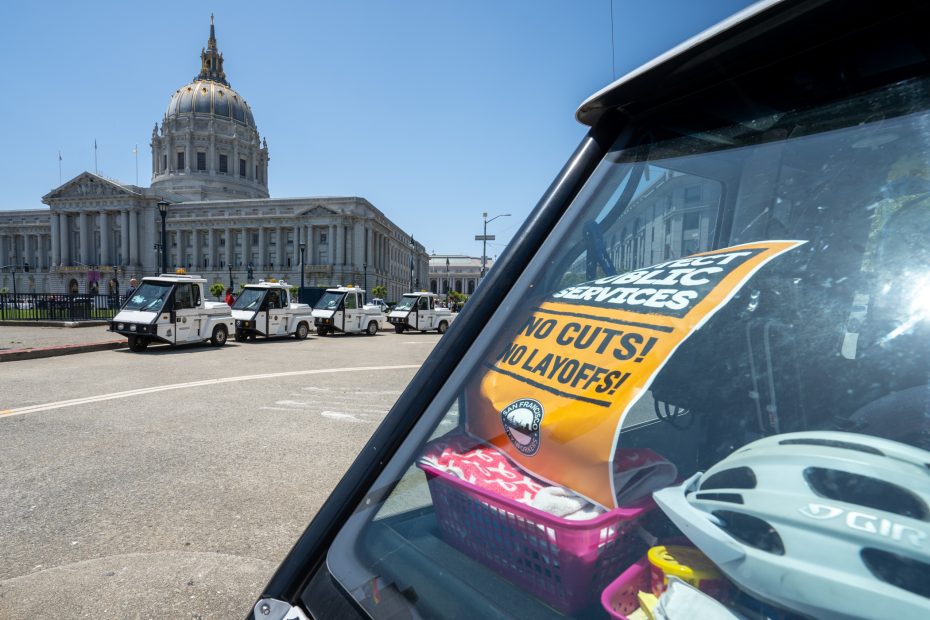 A yellow sign reading "Protect Public Services, No Cuts! No Layoffs!" is seen inside a parked vehicle near a government building with a dome. Other service vehicles are lined up outside.