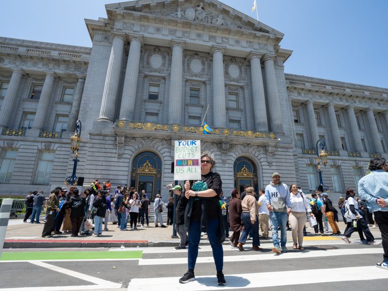 A person holds a sign reading "Your Budget Cuts Hurt Us All" in front of a large government building, surrounded by a crowd of people.