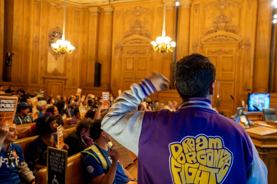 A person in a purple jacket reading "Dream Organize Fight" stands before a seated audience in a wood-paneled room, raising their fist as others hold up signs.