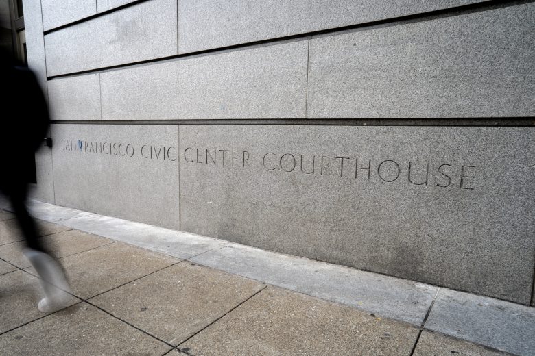 A person walks past a concrete wall with the inscription "San Francisco Civic Center Courthouse" on a city sidewalk.