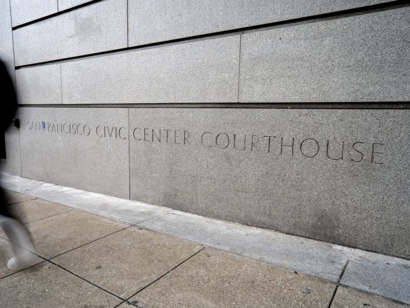 A person walks past a concrete wall with the inscription "San Francisco Civic Center Courthouse" on a city sidewalk.
