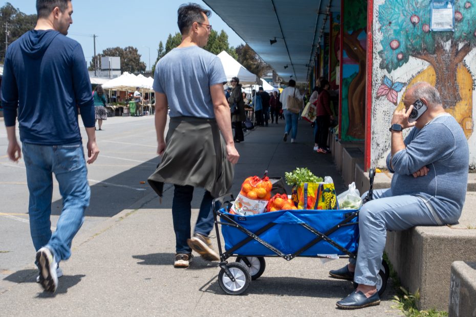 A man sits on a low wall talking on the phone beside a blue wagon filled with groceries, while pedestrians walk by on a sidewalk near outdoor market stalls.