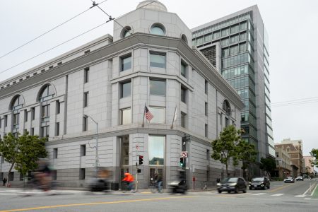 A gray stone building with an American flag stands on a street corner, with blurred cars and cyclists passing by in the foreground.