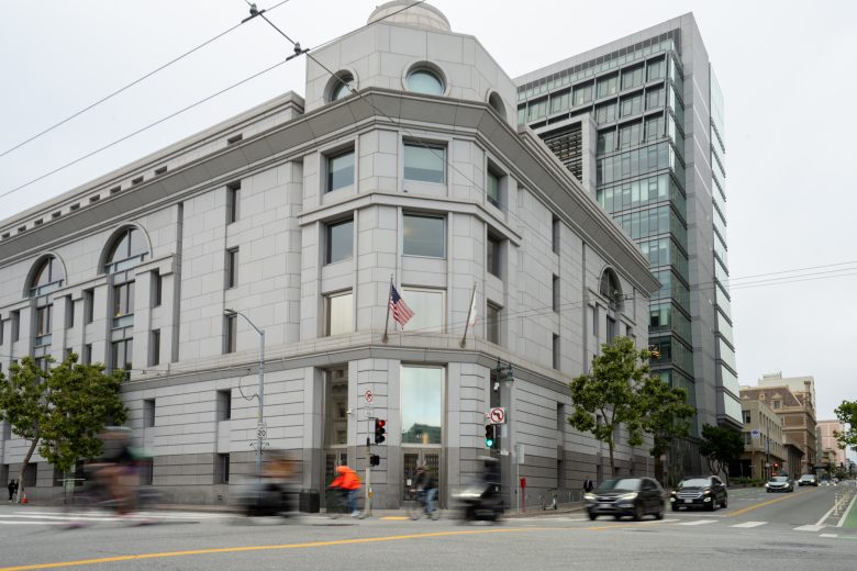 A gray stone building with an American flag stands on a street corner, with blurred cars and cyclists passing by in the foreground.