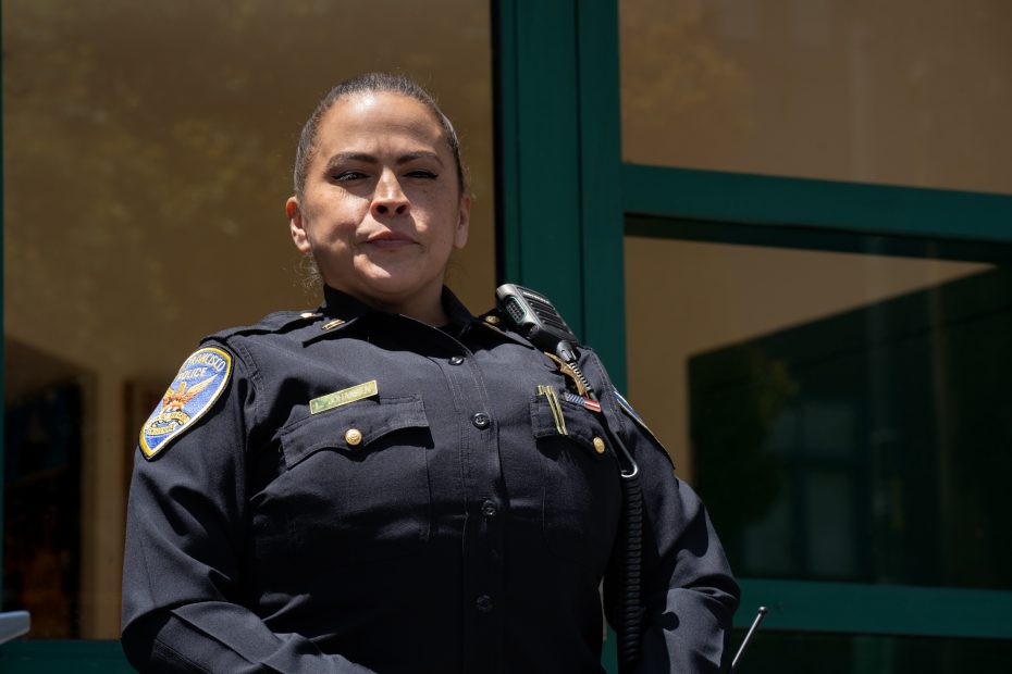 A police officer in uniform stands outdoors in front of a building, looking slightly upward with a serious expression.