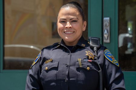 A police officer in uniform stands in front of a green door, smiling at the camera.