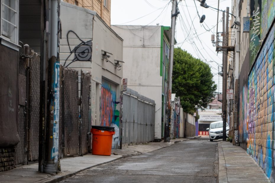 Narrow urban alley with graffiti on walls, a bright orange trash bin, metal fences, overhead wires, and a white car parked near the end of the alley.