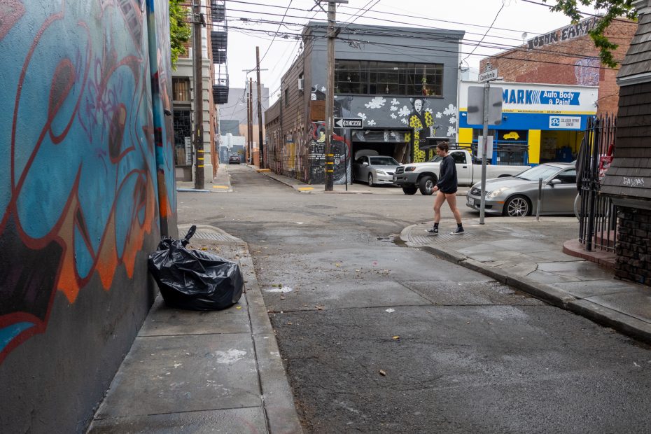 A narrow urban alleyway with graffiti on the left wall, a garbage bag on the sidewalk, parked cars, and a person crossing the street.