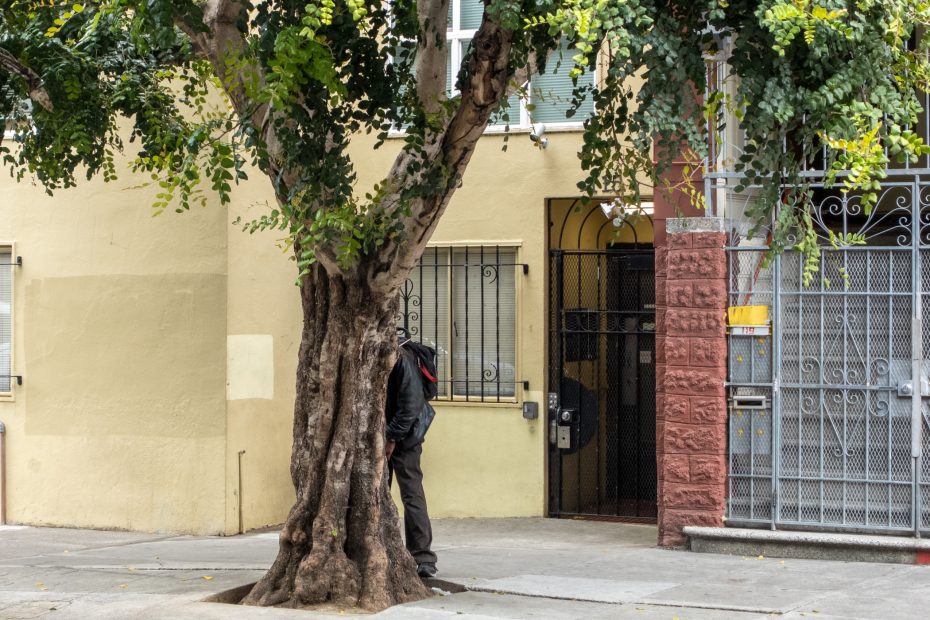 A person stands partially hidden behind a large tree on a city sidewalk in front of a yellow building and a gated entrance.