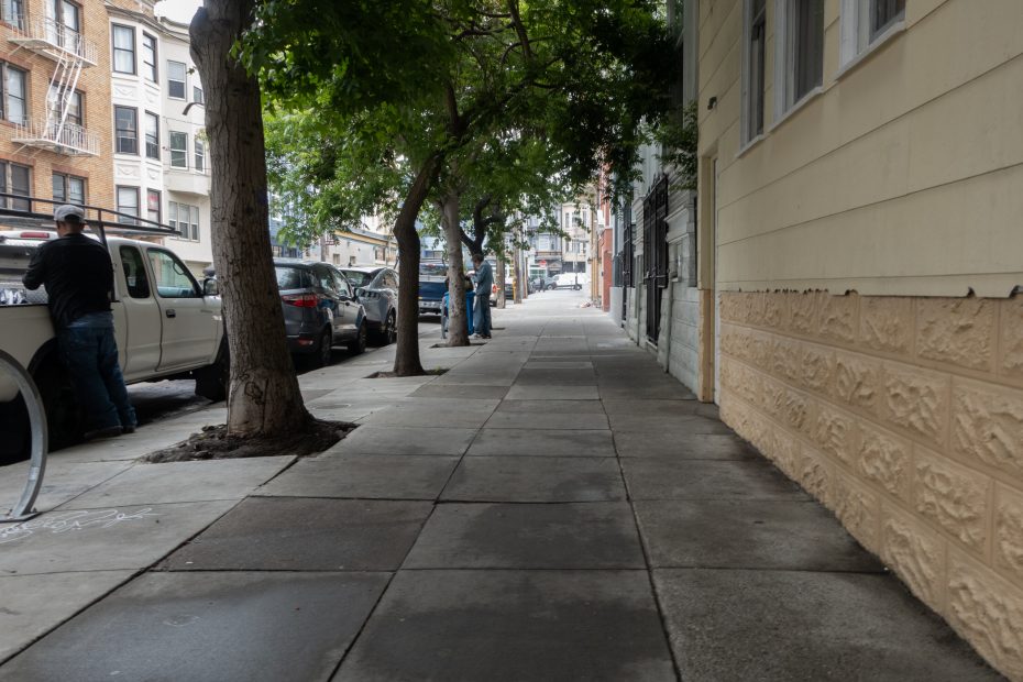 A city sidewalk lined with parked cars and trees, with a few people standing by vehicles and buildings on either side.